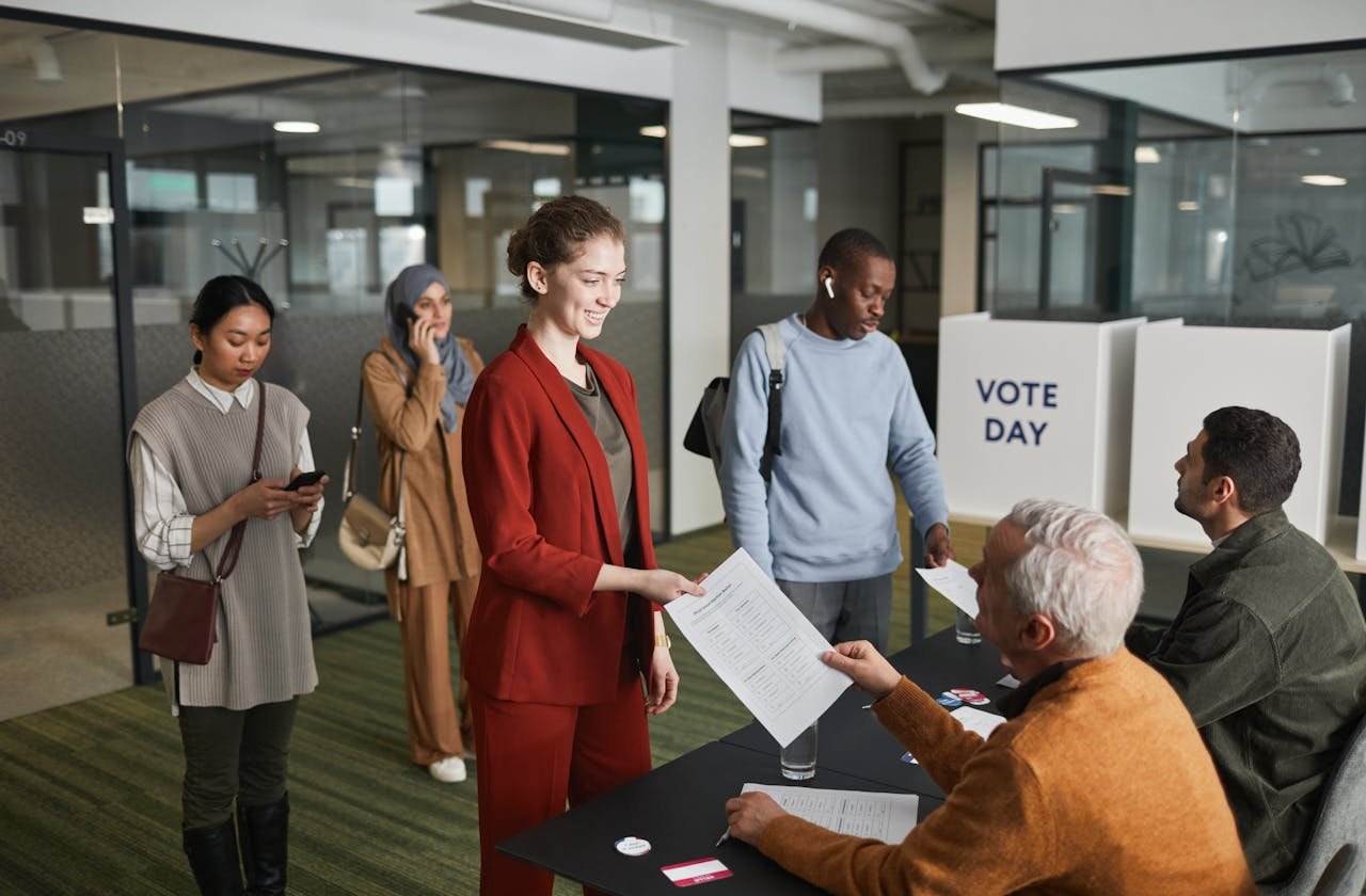 A diverse group of adults participating in an indoor voting process, engaging with election officials.